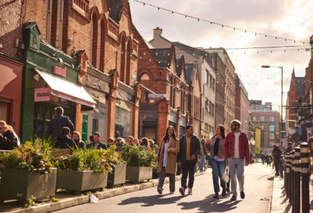 A cheerful group strolls along a lively street with historic brick buildings and outdoor cafés.