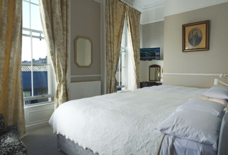 Elegant guest room with large bed, cosy furnishings, and sunlight through the curtains.