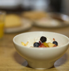 A bowl of fresh fruit and yoghurt on a wooden table, with orange juice in the background.