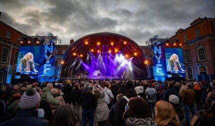 A lively crowd enjoys an outdoor concert, with a musician performing on a vibrant lit stage.